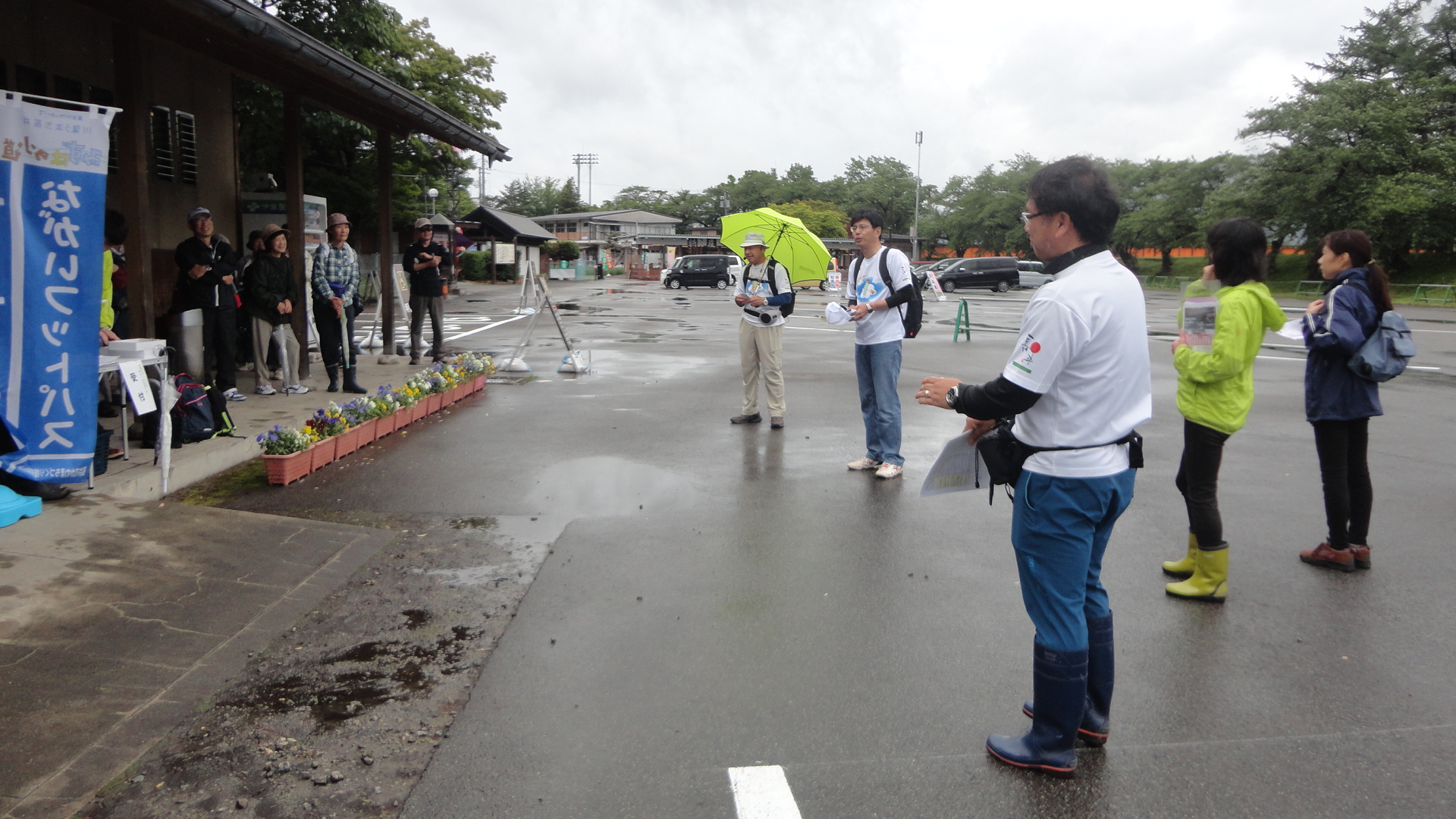 小雨が降る中、あやめ公園の駐車場に参加者の方々が集まり、開会式が行われている様子の写真