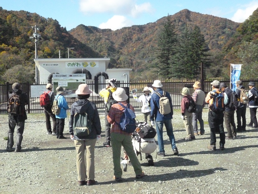 柵に囲まれた場所に小水力発電所があり、参加者の方々が発電所前に集まって見学をしている様子の写真