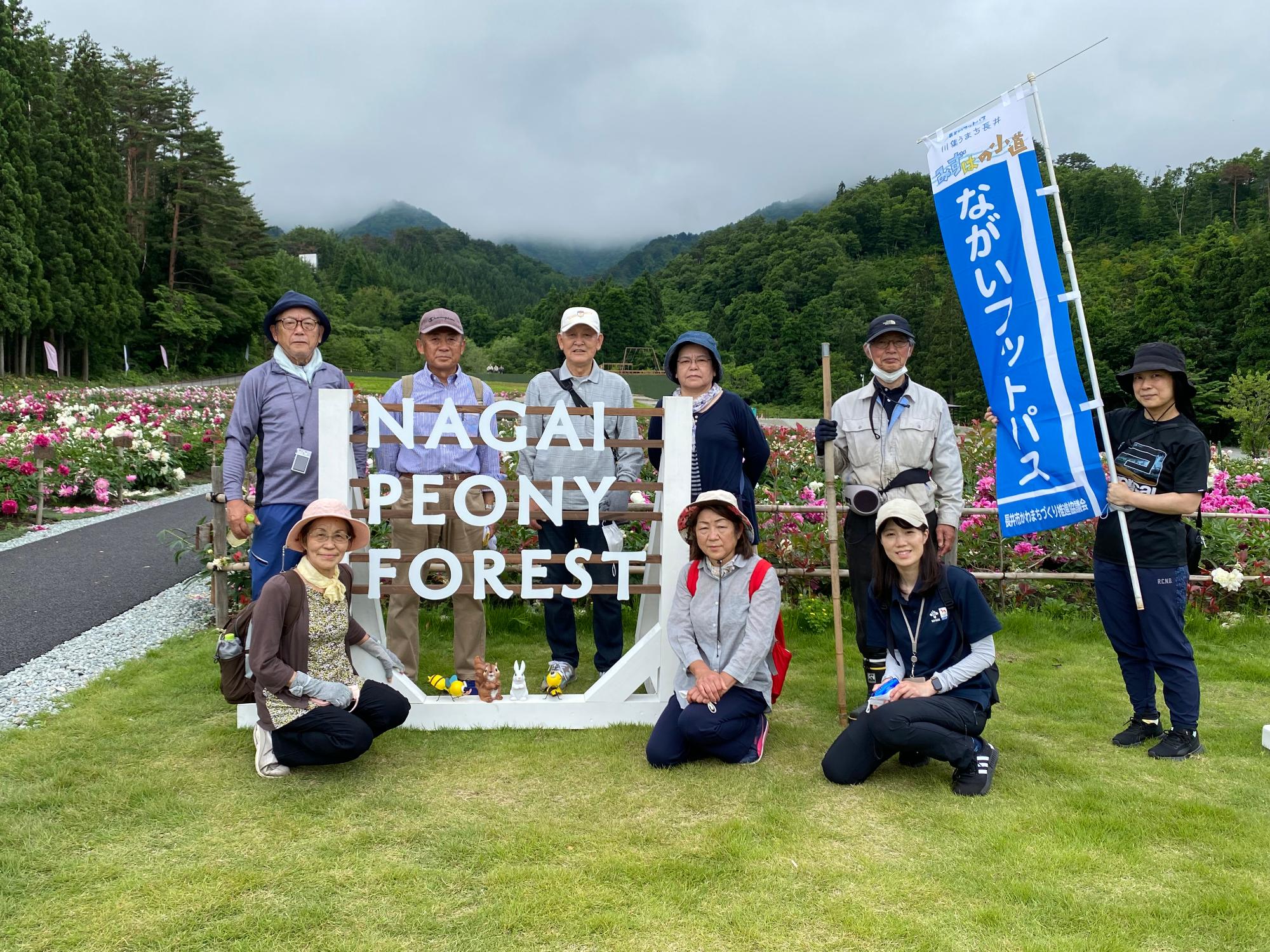 参加者の方々が「ながいフィットパス」と書かれたのぼり旗を持ち、「NAGAI PEONY FOREST」と書かれた看板の周りに集まり、写っている集合写真