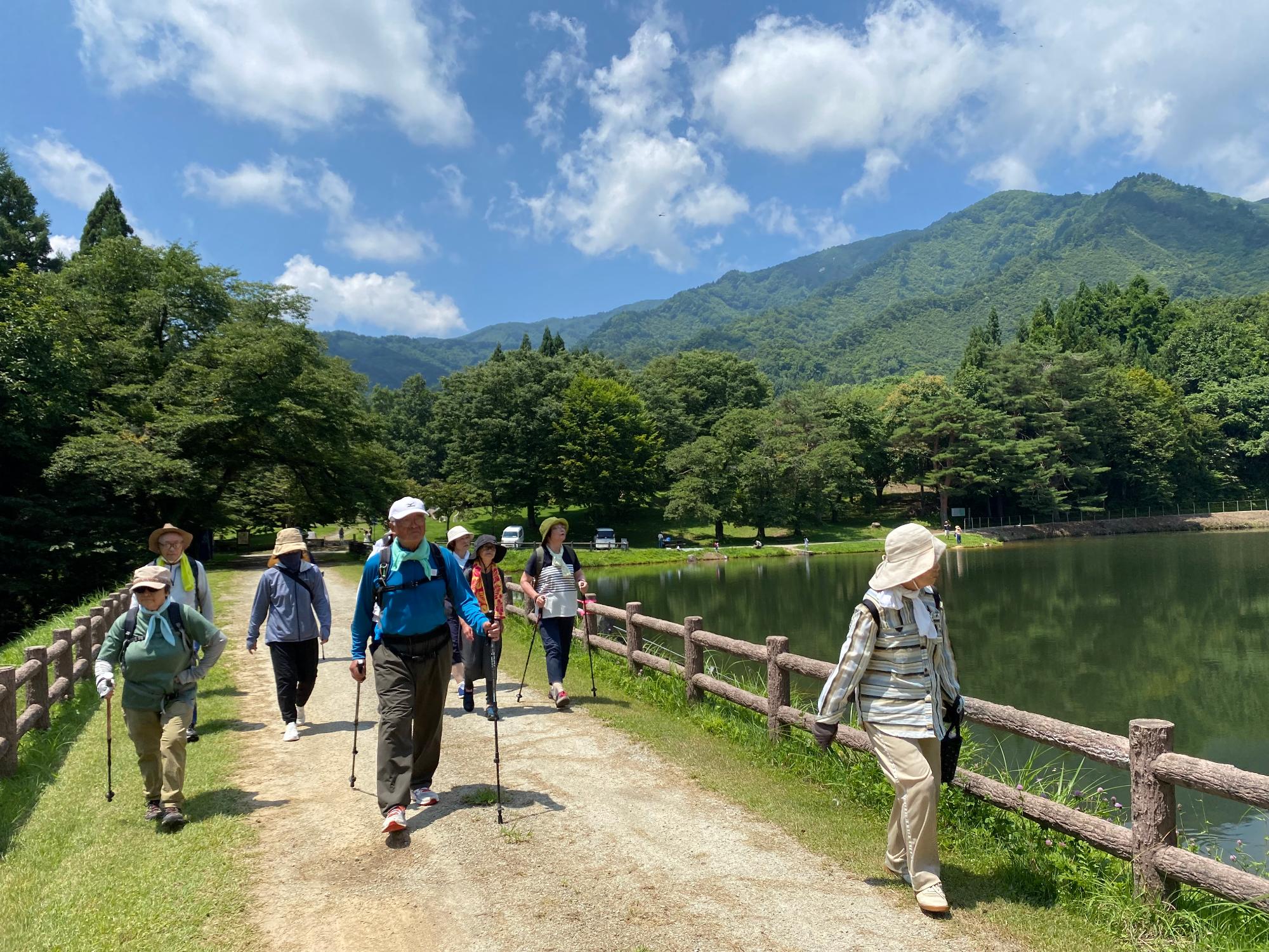 青空に白い雲が浮かび、後方には山並みと緑の木々が広がる中里堤の湖側を参加者の方々が歩いている様子の写真