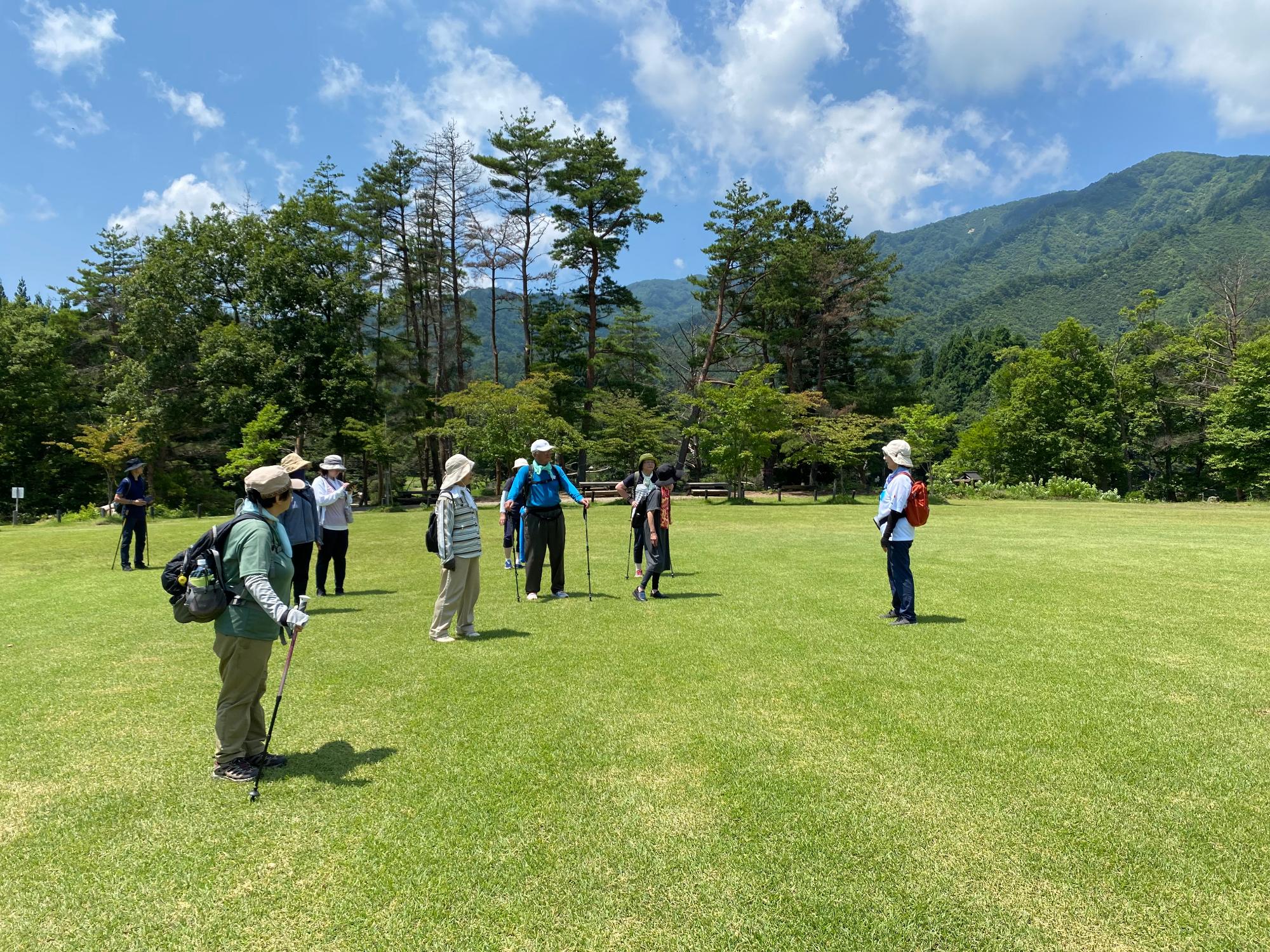 青空に白い雲が浮かび、後方には山並みと緑の木々が見え、芝生の広々とした太陽の広場に参加者の方々が集まっている写真