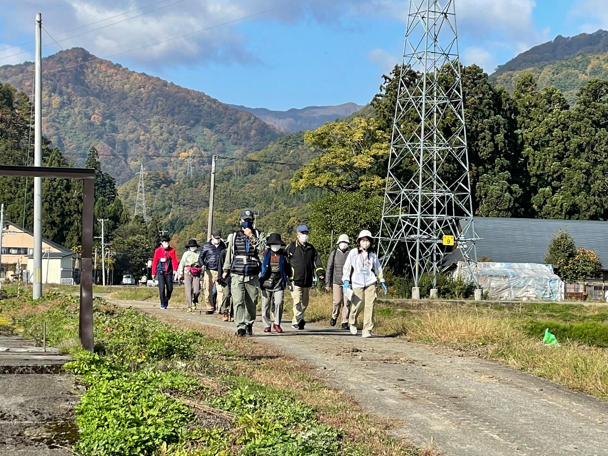 後方には山並みと木々が見え、周りには田畑が広がるのどかな田園風景の中を参加者の方々が歩いている様子の写真