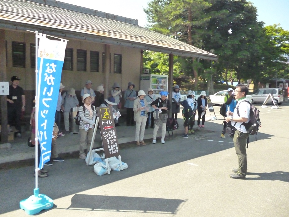 あやめ公園の駐車場にある建物の軒下に参加者の方々が集まり、主催関係者と思われる男性の話を聞いている開会式の様子の写真