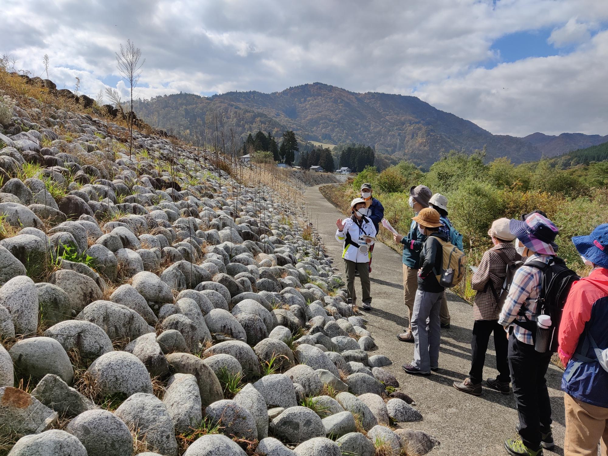 沢山の石が積み上げられて造られた平山締切堤防前で、参加者の方々がガイドさんの説明を聞いている様子の写真